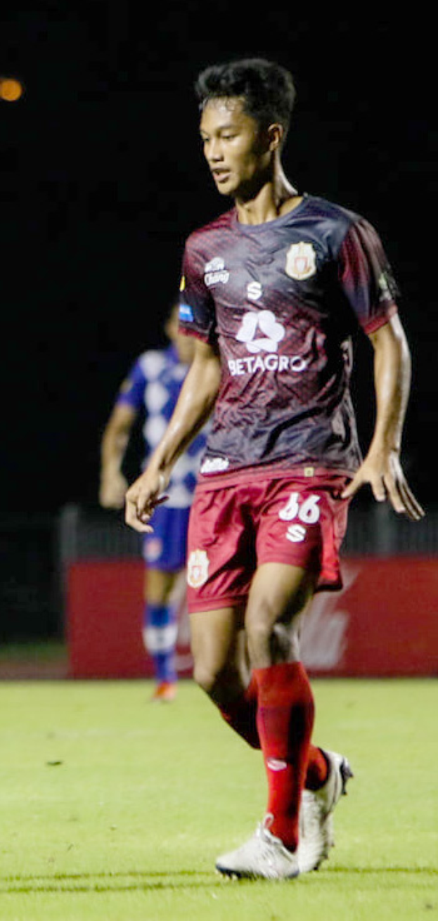Ein Mann in einem maronenfarbenen Hemd und roten Shorts spielt Fußball auf einem Feld, mit einer anderen Person im Hintergrund und einer Tafel mit Text.