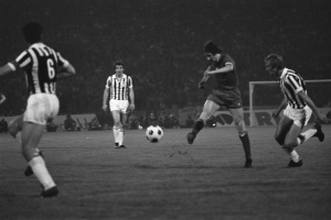 Eine Gruppe von Männern, die auf einem Feld Fußball spielen, mit einem Tor im Hintergrund und Zuschauern, die von der Seitenlinie aus zusehen, dargestellt in Schwarz-Weiß.