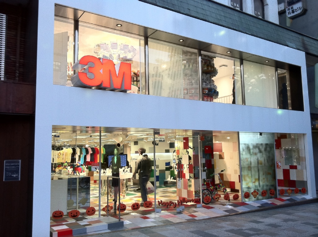 Ein Geb√§ude mit einer Glaswand, die Kleider auf B√ľgeln, eine Schaufensterpuppe, ein Fahrrad, aufgeklebte Bilder und auf die Scheibe geschriebene Texte zeigt.