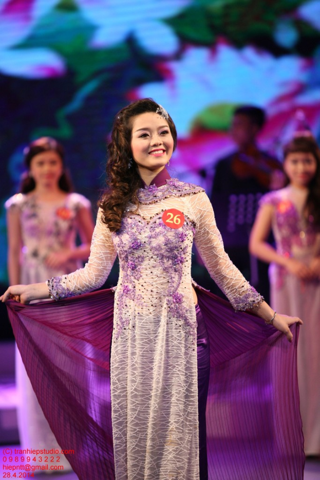 Eine Frau in einem cremefarbenen und lila Kleid mit einem Anstecker daran, vor einem unscharfen Hintergrund.