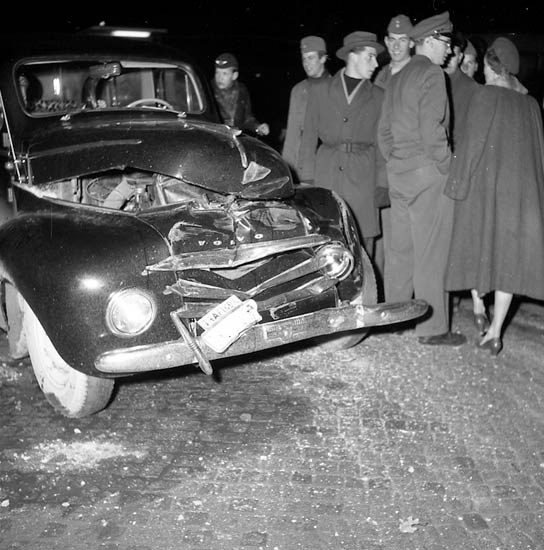 Schwarz-weißes Bild eines schwerbeschädigten Autos am Straßenrand mit einer Gruppe von Menschen, die es inspizieren.