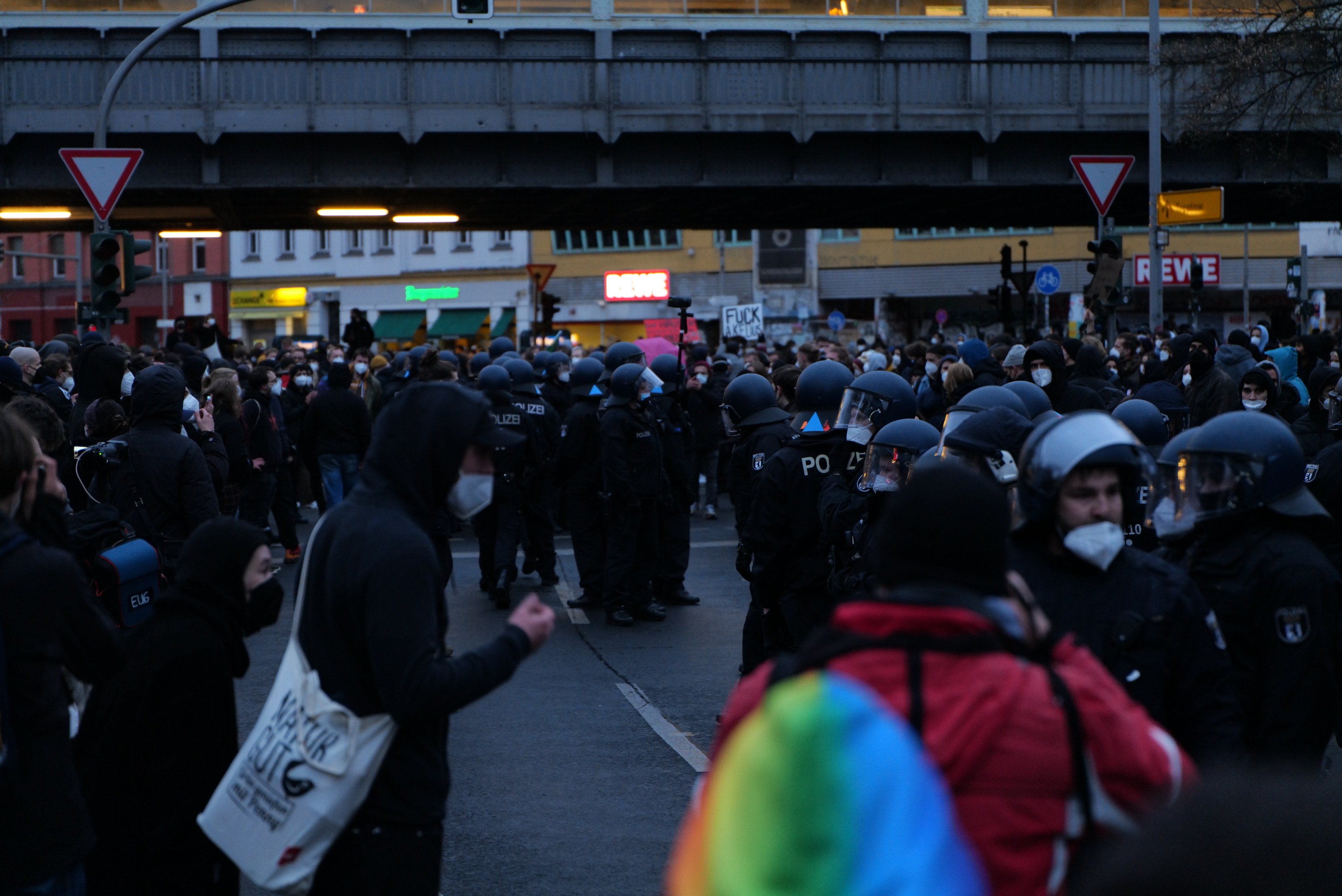 Große Gruppe von Menschen vor einer Reihe von Polizeibeamten, einige mit Helmen und Taschen oder Kameras, mit Gebäuden, Schildern, Verkehrsampeln, einer Brücke und einem Baum im Hintergrund.