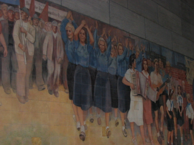 Eine Gruppe von Menschen versammelte sich, mit blauen-kleidsenden Mädchen in der Mitte, die klatschten, Musikern, die Instrumente spielten, Kindern, die in der rechten Ecke spielten, und Individuen, die rote Fahnen in der linken Ecke hielten.