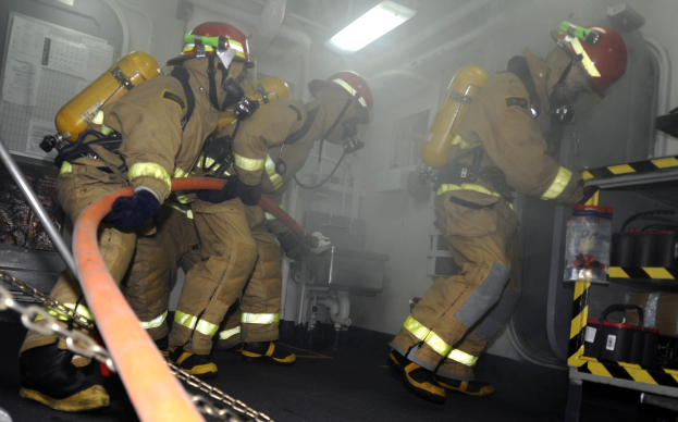 Feuerwehrleute in Schutzausrüstung und Helmen arbeiten an einem Feuerwehren, mit Equipment-Regalen und Hintergrund-Infrastruktur sichtbar.