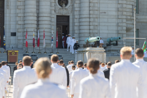 Gruppe von Menschen in weißer Marineuniform, die auf einer Treppe vor einem Gebäude mit Säulen und einer Tür stehen, bei einer Abschlussfeier mit Fahnen, einem Podium und Kanonen im Hintergrund.