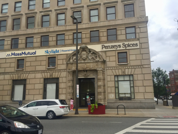 Großes Gebäude mit parkenden Autos, Straßenlaternen, Lichtern, fahrenden Fahrzeugen, Fußgängern, Grünflächen und bewölktem Himmel, identifiziert als Hauptsitz der Massachusetts Mutual Financial Group.