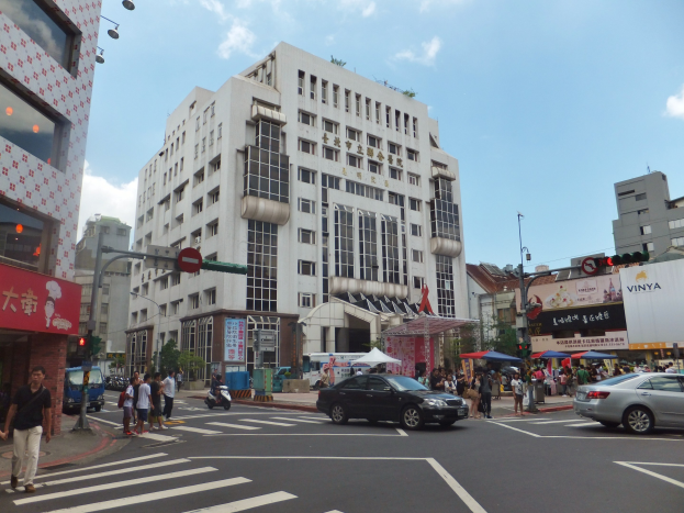 Eine belebte Stadtstraße mit Fußgängern, Fahrzeugen und hohen Gebäuden im Hintergrund bei bewölktem Himmel.
