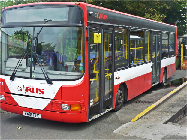 Ein roter Stadtbus mit weißen Linien, der eine Windschutzscheibe, Glasfenster und Türen hat und auf einer Straße mit Bäumen im Hintergrund fährt.