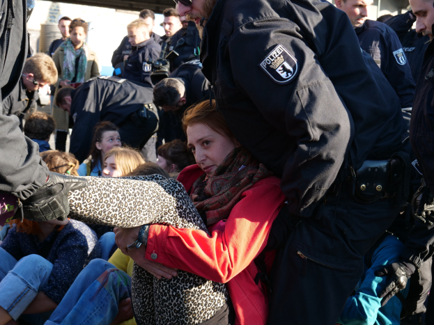 Eine Frau wird von einem Polizeibeamten vor einer Menge eskortiert, wobei einige Menschen sitzen und andere stehen, ein Schuppen und ein klarer blauer Himmel im Hintergrund.