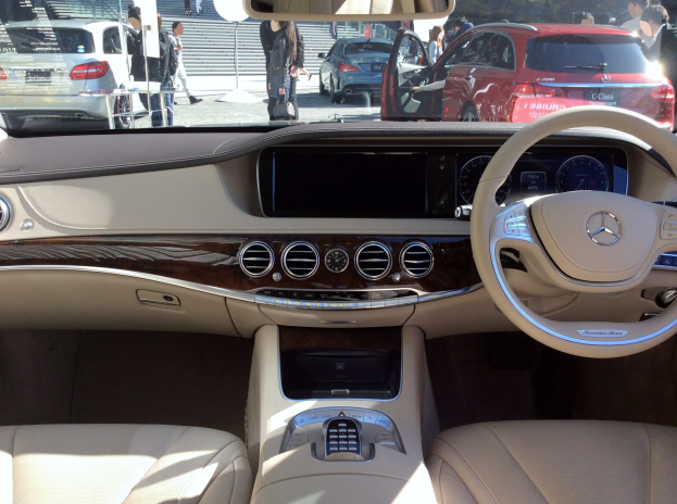 Das Innere eines 2016 Mercedes-Benz S-Klasse, mit einem Lenkrad, Armaturenbrett, Knöpfen und Sitzen, mit einer Sicht auf andere Fahrzeuge, Fußgänger, Bäume und ein Gebäude außerhalb.