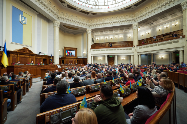 Eine große Gruppe von Menschen in einem großen Raum mit Flaggen, Tischen mit Mikrofonen, einer Wand mit einem Bildschirm, Säulen, Deckenleuchten und einigen stehenden Personen, die eine Sitzung des ukrainischen Parlaments darstellen.