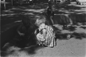 Ein Schwarz-Weiß-Foto von drei Kindern, die im Sand spielen, mit einem Jungen und einem Mädchen, die neben einer Schaufel auf dem Boden sitzen, und ein paar Menschen, die auf Stählen sitzen, mit einem Wagen rechts daneben.