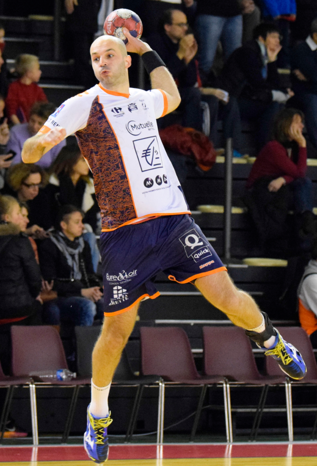 Mann in der Luft mit einem Handball, umgeben von Menschen auf Stühlen und einigen im Hintergrund Stehenden