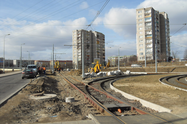 Baustelle mit einer durchfahrenden Bahnschiene, umgeben von Geb├Ąuden, Stra├čeninfrastruktur, Kraftfahrzeugen, Personen, Schutt, B├Ąumen und einem bew├Âlktem Himmel.