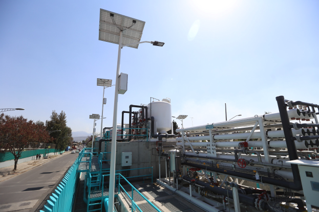 Großes industrielles Gebäude mit ausgedehnten Rohrleitungen und Solarpanelen, umgeben von Sicherheitsgeländern und Infrastruktur, vor einem klaren blauen Himmel auf einem Hügel, Teil einer California-Wasseraufbereitungsanlage.