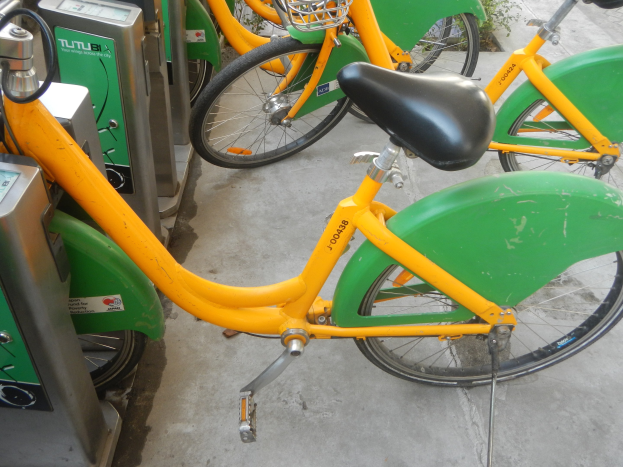 Eine Reihe gelber und grüner Fahrräder nebeneinander auf dem Boden geparkt, mit Maschinen links und Pflanzen im Hintergrund.