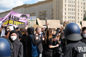 Gruppe von maskierten Menschen mit Schildern vor einem Gebäude, mit zwei Polizisten in Helmen auf der rechten Seite, Bäumen und bewölktem Himmel im Hintergrund.