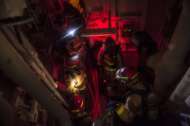 Feuerwehrleute in Helmen und Sauerstoffflaschen stehen in einem schwach beleuchteten Raum mit Rohren auf beiden Seiten, bereit für einen Rettungseinsatz in der Nähe von Maschinen und anderen Gegenständen.