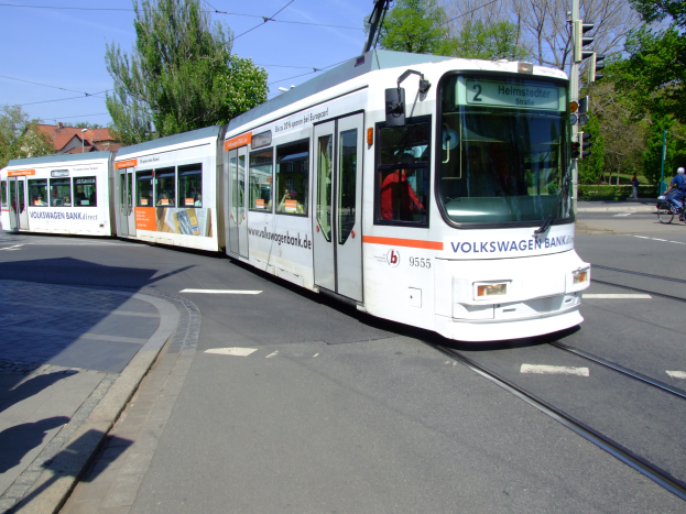 Eine weiße und orangefarbene Straßenbahn befindet sich auf einer Straße in der Stadt mit einem Fahrradfahrer auf der rechten Seite, Bäumen, Gebäuden und einem klaren blauen Himmel im Hintergrund.
