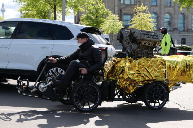 Ein Mann im Rollstuhl mit einem großen Motor daran, umgeben von Fahrzeugen auf einer Straße mit Bäumen, Gebäuden und einem klaren blauen Himmel im Hintergrund.