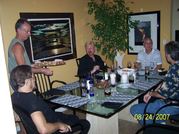 Eine Gruppe von Menschen sitzt an einem Esstisch mit Weingläsern, Flaschen und Schüsseln mit Essen, ein Mann serviert, Fotos auf dem Wandregal und eine Pflanze auf der Seite.