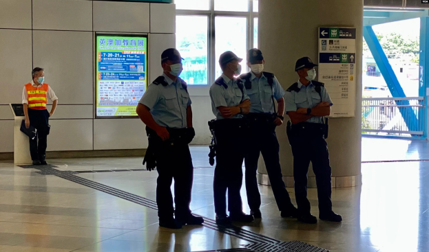 Eine Gruppe von Polizisten in Masken steht vor einem Gebäude mit Glastüren, Schildern mit Text, einer Säule und einem Geländer, mit Bäumen und Fahrzeugen im Hintergrund, was darauf hinweist, dass sie in Hong Kong verhaftet wurden.