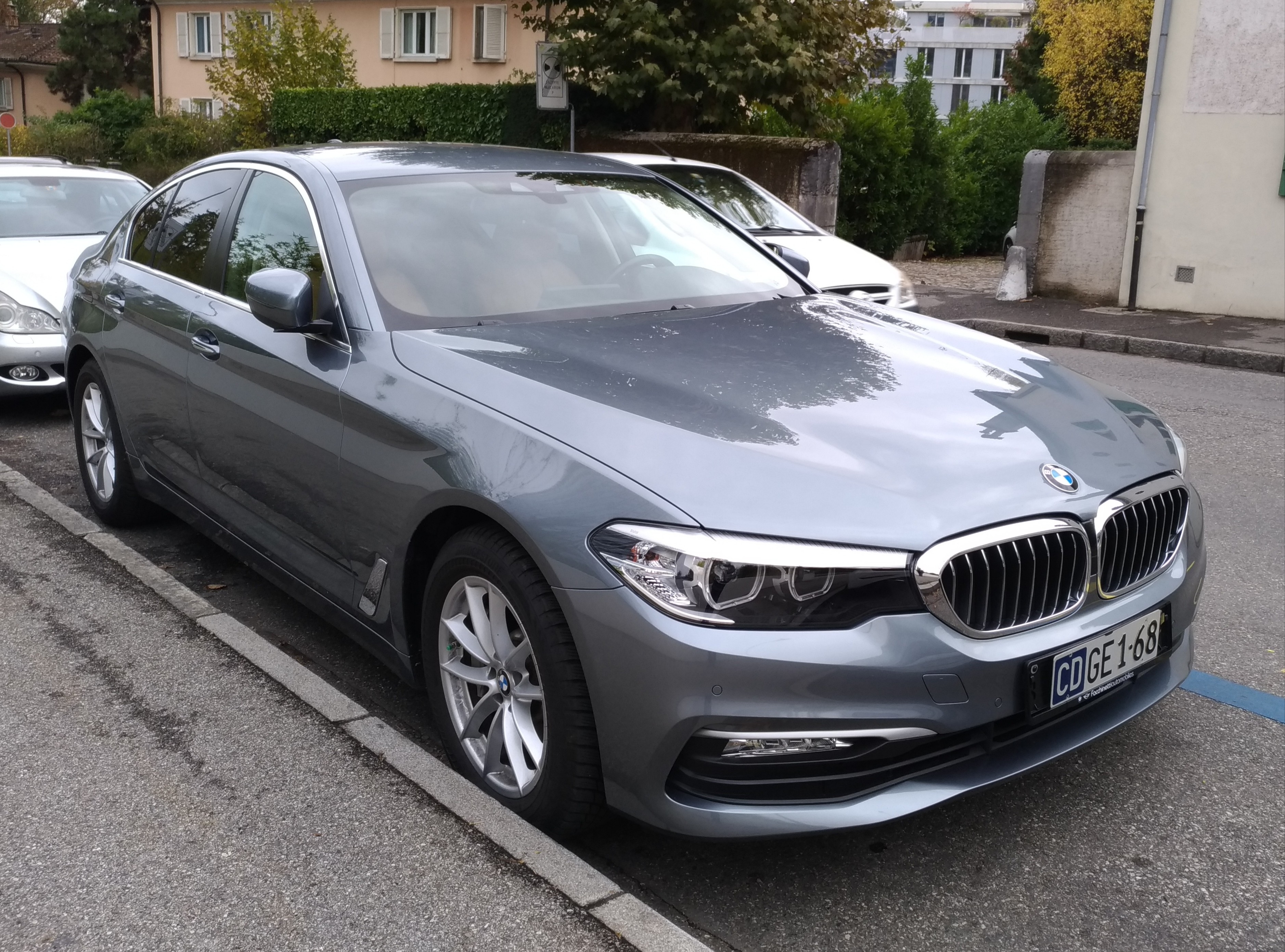 Ein BMW 5er 520d xDrive Luxury Line f√§hrt auf einer Stra√Ÿe mit Geb√§uden, B√§umen, Pflanzen und einem klaren blauen Himmel im Hintergrund.