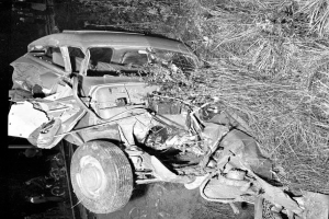 Ein Schwarz-Weiß-Foto eines schwer beschädigten Autos am Straßenrand mit einer zerbeulten Front, zersplitterten Fenstern, einem Baum im Hintergrund und verstreuten Gegenständen.