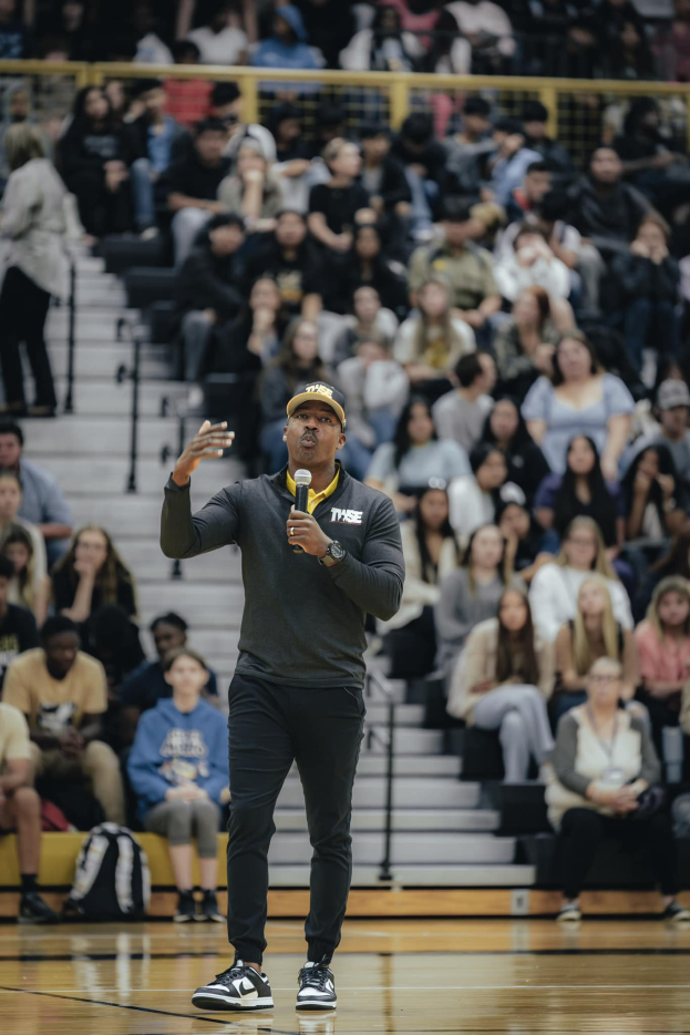 Ein Mann mit Mütze hält ein Mikrofon auf einem Basketballfeld, mit einer Gruppe von Menschen auf einer Treppe und Taschen auf dem Boden hinter ihm.