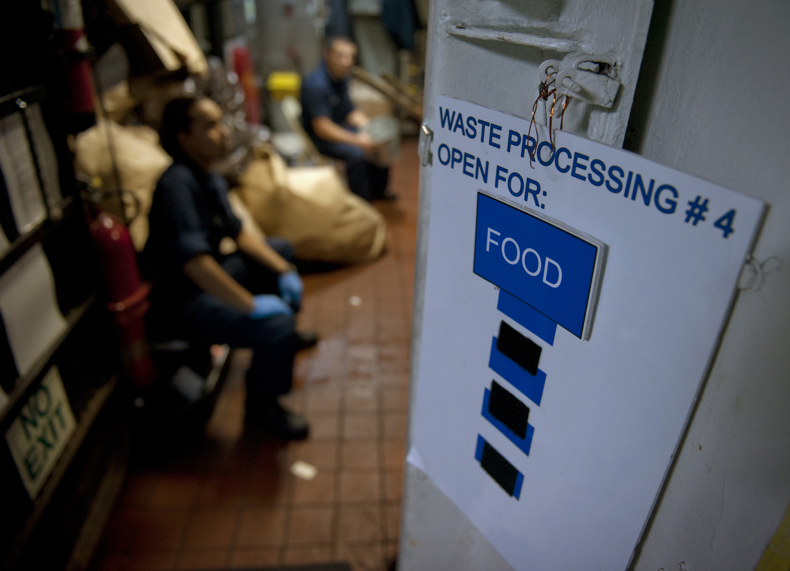 Ein Mann sitzt auf dem Boden neben einem Schild mit der Aufschrift "Abfallverarbeitung 4: Open for Food", mit einer Tür auf der rechten Seite, Regalen mit Gegenständen auf der linken Seite und unscharfen Figuren und Taschen im Hintergrund.