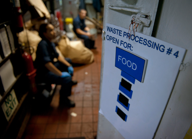 Ein Mann sitzt auf dem Boden neben einem Schild mit der Aufschrift "Abfallverarbeitung 4: Open for Food", mit einer Tür auf der rechten Seite, Regalen mit Gegenständen auf der linken Seite und unscharfen Figuren und Taschen im Hintergrund.