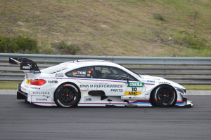 Ein BMW M4 DTM-Rennwagen auf einer Rennstrecke, mit Text auf dem Wagen und einer Begrenzung im Hintergrund, umgeben von saftigem Grün.