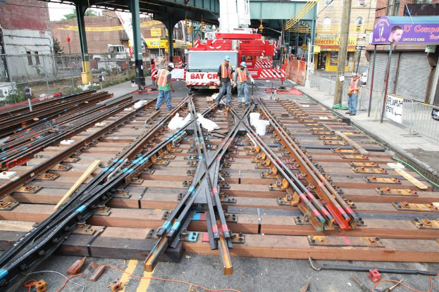 Arbeiter in Jacken und Mützen bauen Schienen, mit einem Fahrzeug mit Materialien beladen und einem Schuppen mit Pfosten im Hintergrund und einem Geschäft auf der anderen Seite.