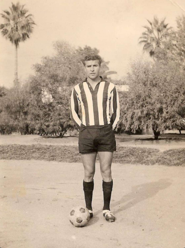 Ein Schwarz-Weiß-Foto eines älteren Mannes mit einem Fußball, der draußen vor Bäumen und Himmel steht.