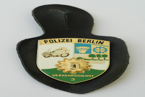 Ein Polizeistern mit der Aufschrift "Polizei Berlin", der ein blaues Schild mit einem weißen Stern in der Mitte zeigt, umgeben von einem weißen Rand, auf einer Oberfläche platziert.