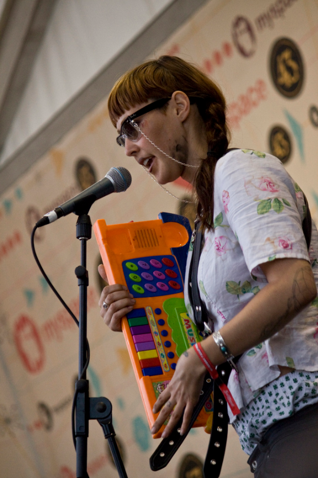 Eine Frau in einem wei├čen Hemd und Sonnenbrille singt in ein Mikrofon, waehrend sie ein Piano spielt, mit einem Plakat an der Wand dahinter.