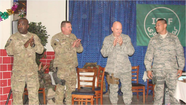 Vier Personen stehen, drei klatschen, eine sitzt auf einem Stuhl neben einer Pflanze, mit einem Banner an einem Vorhang im Hintergrund, alle in militärischer Kleidung, mit Stühlen und einem Tisch im Sichtbereich.