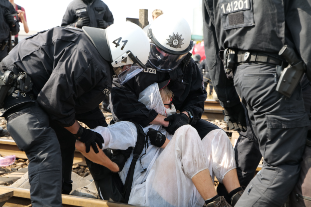 Eine Gruppe von Polizisten in schwarzer Einsatzkleidung verhaftet einen Mann in einem weißen Kleid auf einem Bahnsteig, mit anderen Menschen und dem Himmel im Hintergrund.