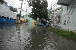 Eine überflutete Straße mit einem Motorrad in der Mitte, umgeben von Gebäuden, Bäumen und Strommasten mit Drähten, mit ein paar Menschen und Fahrzeugen im Hintergrund.