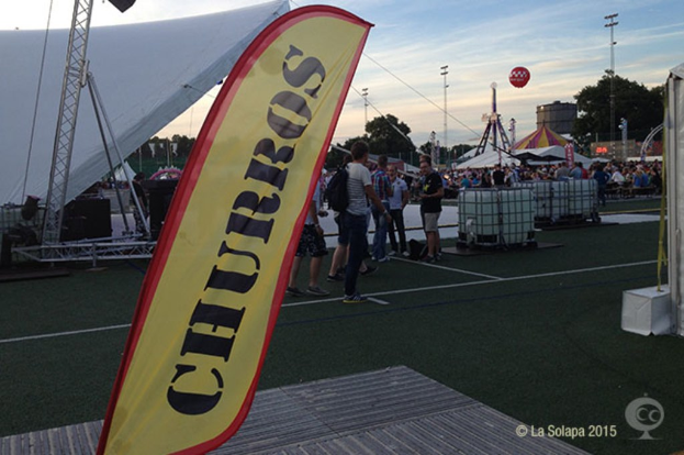Eine volle KarnevalsSzene mit Zelten, einem zentralen Mast, Anhängern auf der Straße, Bäumen, Strommasten und einem eisernen Gestell im Hintergrund unter einem Himmel.