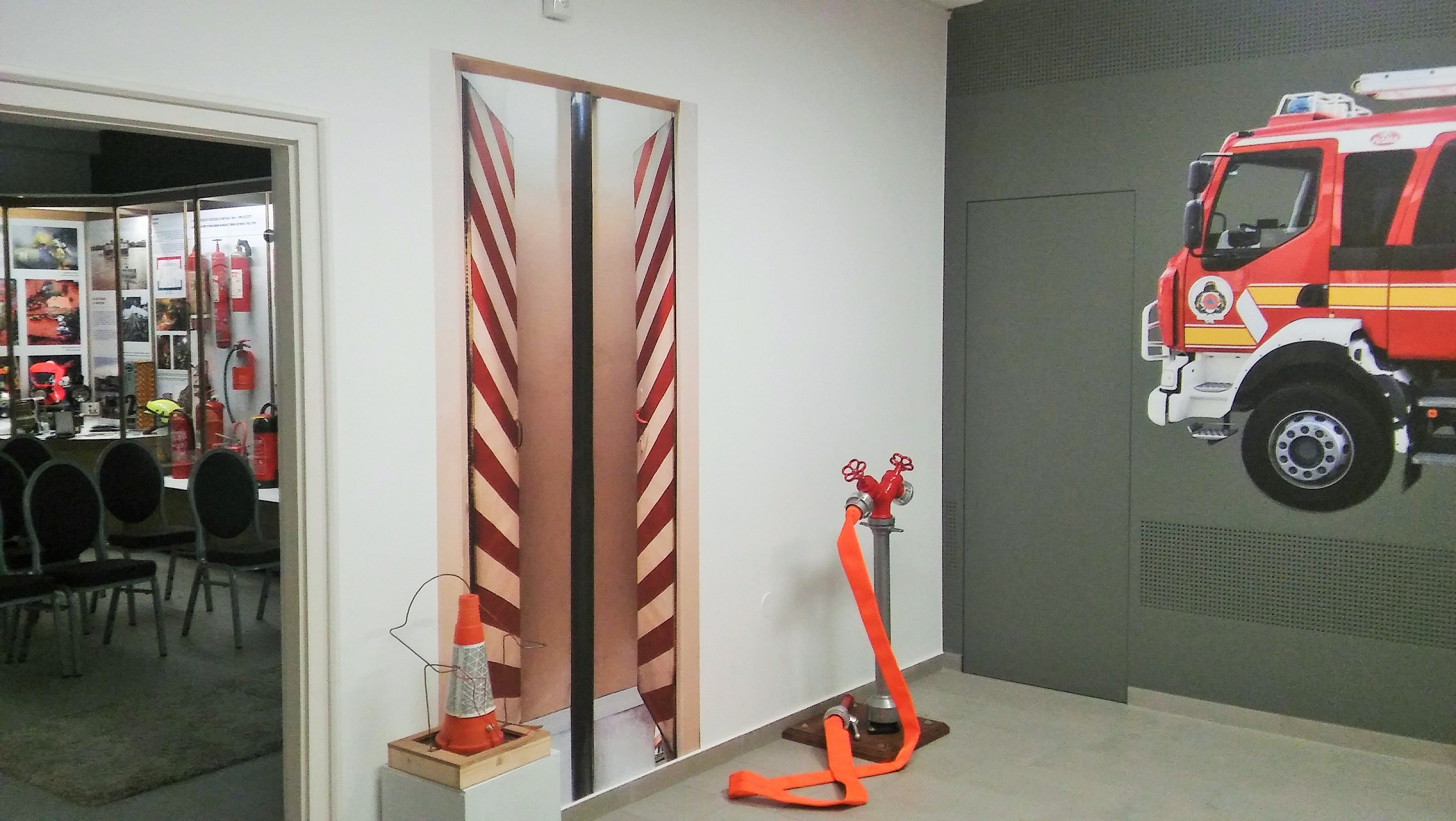 Zimmer mit einer Wandmalerei eines Feuerwehrautos, einer Tür, einem Verkehrskegel, einem Ständer, einem Teppichboden, Stühlen, Tischen und Feuerlöschern.