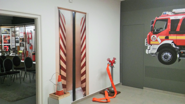 Zimmer mit einer Wandmalerei eines Feuerwehrautos, einer Tür, einem Verkehrskegel, einem Ständer, einem Teppichboden, Stühlen, Tischen und Feuerlöschern.