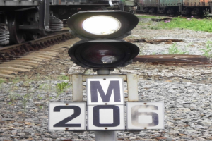 Eine Bahnübergangssignalbox auf einem Pfahl mit einem Zug auf den Schienen im Hintergrund, umgeben von Steinen und Gras.