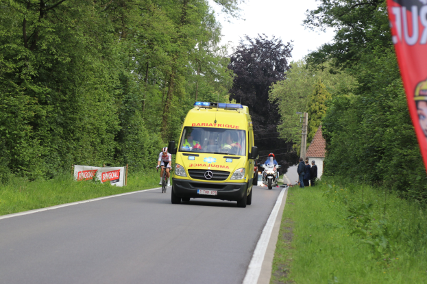 Ambulanz fährt auf einer Straße mit Radfahrern äquidistant, Gras und Bäume auf beiden Seiten, Häuser, Mäste und einen klaren blauen Himmel im Hintergrund.