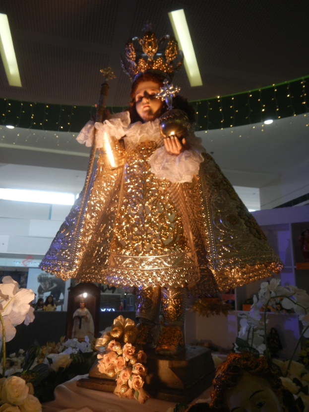 Eine goldbekleidete Statue einer gekrönten Frau, geschmückt mit Blumen und Gegenständen, die auf einem Tisch mit Rahmenfotos und Deckenleuchten im Hintergrund präsentiert wird.