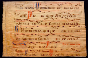 Ein illuminiertes Manuskript mit Musiknotation auf einem Blatt Papier vor einem dunklen Hintergrund, gefüllt mit Text und Musiksymbolen, die ein komplexes Design ergeben.