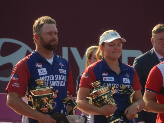 Eine Gruppe von Menschen, die Pokale halten, mit einem "Weltarcherie-Meisterschaften"-Schild im Hintergrund.