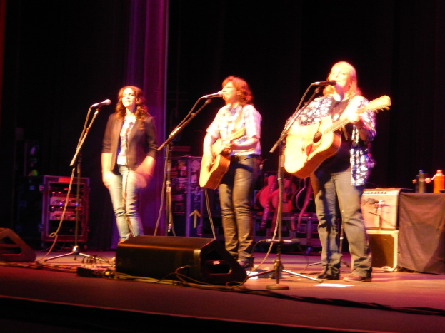 Drei Frauen performen auf der Bühne eines Musikkonzerts, jede singt in ein Mikrofon und spielt eine Gitarre, mit einem Tisch voller Wasserflaschen und Gitarren dahinter.
