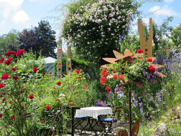 Ein Garten auf der Chelsea Flower Show mit bunten Blumen und Pflanzen, einem Tisch mit einer Decke und Stühlen in der Mitte und Bäumen im Hintergrund vor einem bewölkten Himmel.