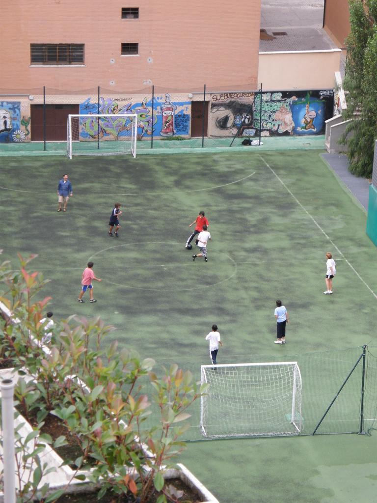 Kinder beim Fußballspielen auf einem Feld mit Toren, einem Gebäude mit Graffiti im Hintergrund, Pflanzen links und Bäumen rechts.